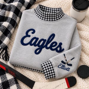 Puede incluir: Una sudadera gris jaspeado con cuello alto de patrón de cuadros vichy en blanco y negro. La palabra Eagles está bordada en cursiva azul marino en el pecho. El puño de la manga presenta un gráfico de palo de hockey azul marino y el nombre Elliott.
