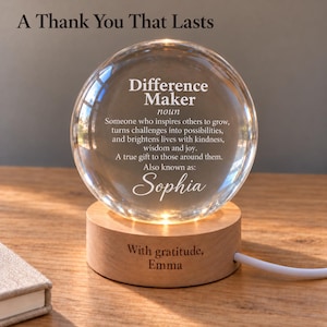 Può includere: Una sfera di vetro trasparente poggiata su una base cilindrica in legno chiaro. La sfera riporta il testo: A Thank You That Lasts, Difference Maker, noun, Someone who inspires others to grow, turns challenges into possibilities, and brightens lives with kindness, wisdom and joy. A true gift to those around them. Also known as: Sophia. La base in legno è incisa con: With gratitude, Emma.