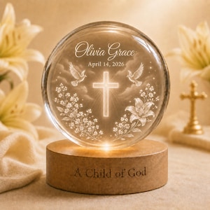Könnte beinhalten: Eine kreisförmige Glasplatte auf einem Holzsockel mit eingraviertem Kreuz, zwei Tauben und Blumenmotiven. Der Text lautet Olivia Grace, April 14, 2026, und A Child of God. Die Platte wird von der Basis aus beleuchtet und erzeugt einen warmen Schein.