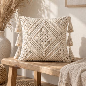 May include: A square off-white throw pillow featuring an intricate macrame knot pattern with a central diamond design. The pillow is accented with three decorative tassels on each side, each adorned with a small, smooth wooden bead at the top.