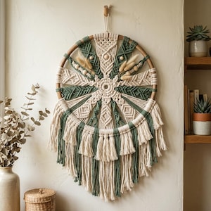 May include: A circular macrame wall hanging featuring a wooden hoop frame with alternating cream and sage green knotted cord patterns. The design includes central wooden beads, dried grass accents, and a layered fringe of hanging cord tassels.