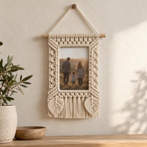 May include: A cream-colored macrame wall hanging frame featuring intricate knotted patterns, a central rectangular photo display, two leaf-shaped fringe accents at the bottom, and a wooden dowel with a hanging cord for wall mounting.