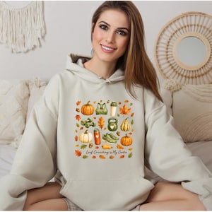 May include: A beige hooded sweatshirt featuring a central graphic print of various autumn-themed items including pumpkins, fall leaves, a kettlebell, dumbbells, sneakers, and a water bottle, with the text Leaf Crunching is My Cardio.