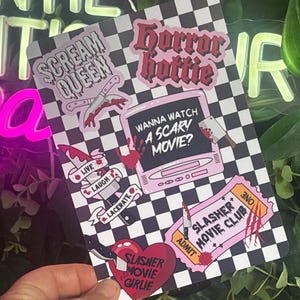 May include: A rectangular sticker sheet with a black and white checkered background featuring horror-themed designs including text that reads Scream Queen, Horror Hottie, Wanna Watch A Scary Movie?, Live Laugh Lacerate, Slasher Movie Club, and Slasher Movie Girlie.