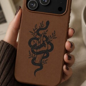 May include: A matte brown smartphone case featuring a black graphic illustration of a hand entwined with a snake and leafy branches. The case has a raised black camera module with three circular lenses and a small circular flash cutout at the top.
