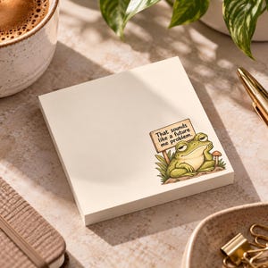 May include: A square, off-white sticky note featuring a printed illustration of a green frog resting its chin on its hand next to a wooden sign that reads, "That sounds like a future me problem." The frog is surrounded by grass and a small mushroom.