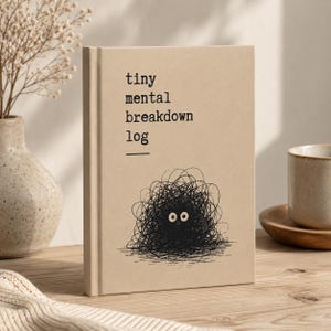 Könnte beinhalten: Ein beigefarbenes Notizbuch mit festem Einband und dem schwarzen Text "tiny mental breakdown log" auf der Vorderseite. Unter dem Text befindet sich eine schwarze Tuschezeichnung eines runden, krakeligen Wesens mit zwei großen, starrenden Augen.