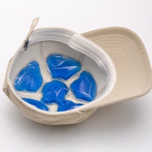 Peut inclure: Une casquette de baseball beige avec une doublure intérieure blanche comprenant cinq inserts de refroidissement en gel bleu disposés en cercle. La casquette est dotée d'un bandeau blanc et d'une sangle réglable avec une boucle en métal.