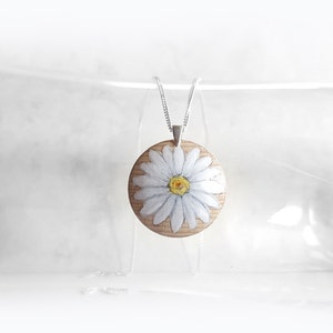 May include: A white daisy painted on a round wooden pendant hangs from a silver chain. The pendant is a light brown colour with a white daisy painted on it. The daisy has a yellow centre and white petals.