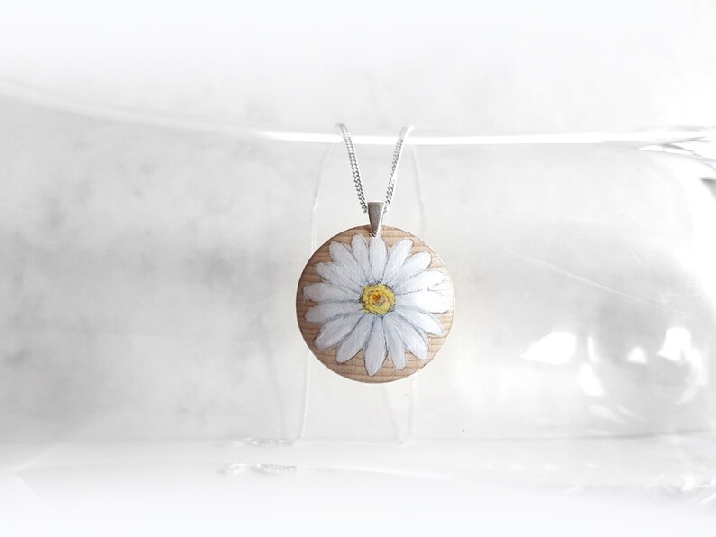 May include: A white daisy painted on a round wooden pendant hangs from a silver chain. The pendant is a light brown colour with a white daisy painted on it. The daisy has a yellow centre and white petals.