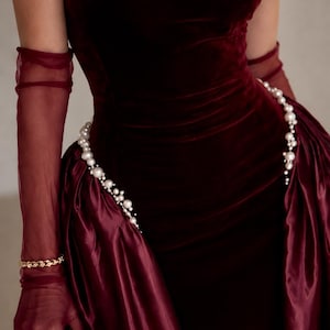 May include: A deep burgundy velvet dress featuring a strapless bodice and a gathered skirt. The waist is accented with a draped string of white pearls. The ensemble includes sheer burgundy opera-length gloves and a gold-toned metallic clutch bag.