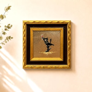 Peut inclure: Une peinture carrée encadrée représentant un chat noir avec des marques blanches tenant un petit verre de vin rouge. Le chat est représenté dans une pose dynamique sur un fond doré texturé. Le cadre est orné, avec une bordure intérieure marron foncé et un bord extérieur en or métallique avec une texture ondulée et festonnée.