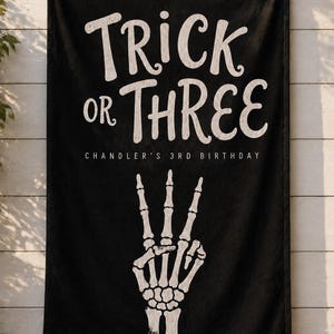 May include: A black rectangular fabric banner featuring white text that reads TRICK OR THREE and CHANDLER'S 3RD BIRTHDAY. Below the text is a white graphic illustration of a skeleton hand making a peace sign gesture with three fingers extended.