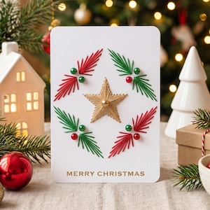 May include: A white rectangular greeting card featuring a central gold star surrounded by four sprigs of red and green thread embroidery. Each sprig is accented with small red and green beads. The text MERRY CHRISTMAS is printed in gold at the bottom.