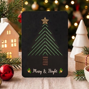 Op de afbeelding: Een rechthoekige zwarte kaart met een geborduurd kerstboomontwerp van groene draad, een gouden ster aan de top en een stam van bruine draad. De tekst Merry & Bright staat in het wit onderaan, geflankeerd door kleine groene hulstblaadjes.