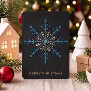 May include: A rectangular black greeting card featuring a central snowflake design embroidered with blue and gold thread. The text MERRY CHRISTMAS is printed in gold at the bottom. The card has rounded corners and a smooth, matte finish.