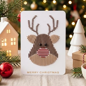 May include: A white rectangular greeting card featuring a reindeer head design created with vertical and horizontal thread stitching in shades of brown, tan, and red. The card has two black bead eyes and the text MERRY CHRISTMAS in gold.