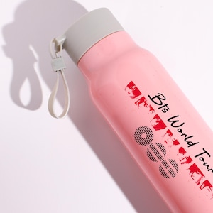 May include: A light pink water bottle with a gray screw-top lid and a matching gray carrying loop. The bottle features black text that reads Bts World Tour along with a series of black circular graphic patterns and small red portrait silhouettes.
