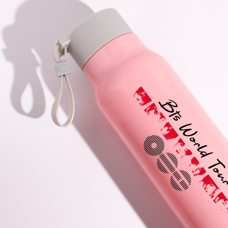 May include: A light pink water bottle with a gray screw-top lid and a matching gray carrying loop. The bottle features black text that reads Bts World Tour along with a series of black circular graphic patterns and small red portrait silhouettes.