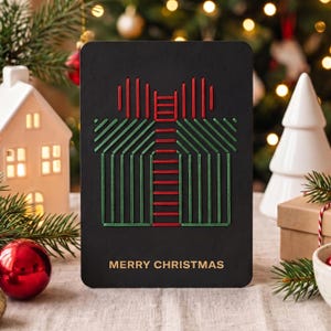 Könnte beinhalten: Eine rechteckige schwarze Grußkarte mit einem stilisierten Geschenkbox-Design aus gesticktem rotem und grünem Garn. Der Text MERRY CHRISTMAS ist in Gold am unteren Rand der Karte aufgedruckt. Das Design besteht aus geometrischen Linien.