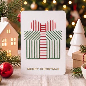 Peut inclure: Une carte de vœux rectangulaire blanche avec un motif brodé de boîte cadeau en fil rouge et vert. Le centre de la carte affiche le texte MERRY CHRISTMAS en lettres dorées. La carte présente des coins arrondis et une surface en papier texturé.