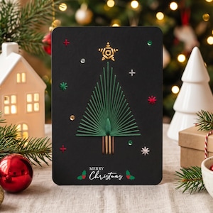 Puede incluir: Una tarjeta de felicitación rectangular negra con un árbol de Navidad de arte con hilos verdes, coronado por una estrella dorada. La tarjeta incluye pequeños detalles bordados, lentejuelas y el texto blanco Merry Christmas con hojas de acebo.