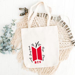 May include: A cream-colored canvas tote bag featuring a red BTS logo graphic in the center. The names RM, Jin, Suga, J-Hope, Jimin, V, and Jung-kook are printed in black text around the red logo, with the word BTS in red at the bottom.