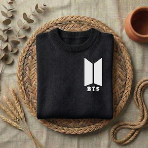 May include: A black long-sleeve sweater featuring a white graphic logo on the chest. The logo consists of two vertical trapezoidal shapes positioned side-by-side with the text BTS printed in white capital letters directly underneath the shapes.