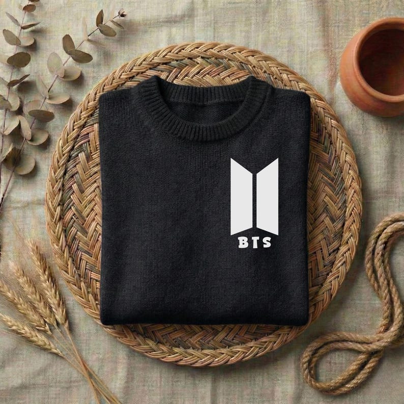 May include: A black long-sleeve sweater featuring a white graphic logo on the chest. The logo consists of two vertical trapezoidal shapes positioned side-by-side with the text BTS printed in white capital letters directly underneath the shapes.
