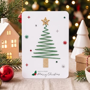 Op de afbeelding: Een witte wenskaart met een groen, met draad geborduurd kerstboomontwerp, een gouden ster als piek en een gouden geborduurde stam. De kaart bevat kleine rode, groene en zilveren accenten en de tekst "HAVE A VERY Merry Christmas".