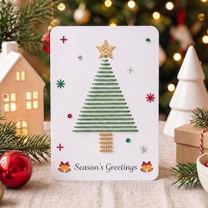 Op de afbeelding: Een witte rechthoekige wenskaart met een geborduurd kerstboomontwerp van groene draad, een gouden ster als piek en een stam van gouden kralen. De kaart bevat de tekst Season's Greetings, kleine rode en groene accenten en twee klokjes.