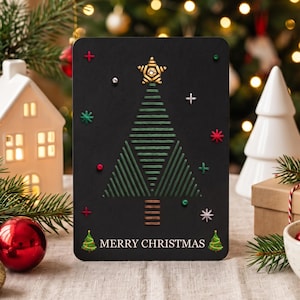 May include: A black rectangular greeting card featuring a stitched green thread Christmas tree design with a gold star topper. The card includes small colourful embroidered accents, silver and red embellishments, and the text MERRY CHRISTMAS.