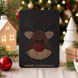 May include: A rectangular black greeting card featuring a stylised reindeer head created with vertical and horizontal thread-like lines in shades of brown, tan, and red. The card has two small black beads for eyes and the text MERRY CHRISTMAS.
