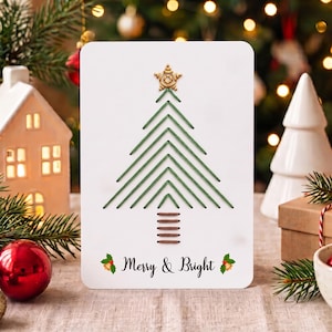 Könnte beinhalten: Eine weiße rechteckige Grußkarte mit einem gestickten Weihnachtsbaum aus grünem Garn, einer goldenen Sternspitze und einem Stamm aus braunem Garn. Der Text Merry & Bright ist in Schwarz unter dem Baum mit kleinen Stechpalmen-Symbolen gedruckt.