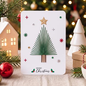Puede incluir: Una tarjeta de felicitación rectangular blanca con un árbol de Navidad de arte con hilos verdes, una estrella dorada en la punta y un tronco de hilo marrón. La tarjeta incluye pequeñas lentejuelas rojas, verdes y plateadas y el texto Merry Christmas.