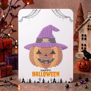 Puede incluir: Una tarjeta rectangular blanca con un diseño de arte con hilos de una calabaza con un sombrero de bruja morado. Sobre la calabaza hay telarañas negras y debajo el texto HAPPY HALLOWEEN en naranja y negro, con una silueta de un pueblo.