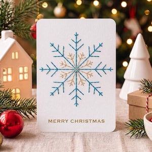 Może przedstawiać: Biała prostokątna kartka okolicznościowa z centralnym wzorem płatka śniegu wyhaftowanym niebieską i złotą nicią. Napis MERRY CHRISTMAS jest nadrukowany złotym kolorem na dole kartki. Kartka ma zaokrąglone rogi i powierzchnię z teksturowanego papieru.