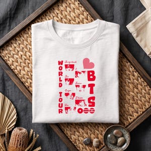 May include: A white crew neck t-shirt featuring a red graphic print. The design includes the text WORLD TOUR and BTS, a heart icon, and small portraits of seven individuals labeled with the names Jung-kook, Jin, Suga, Rm, Jimin, and J-hope.