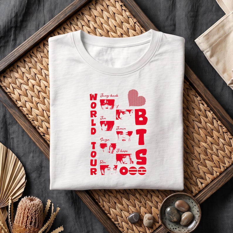 May include: A white crew neck t-shirt featuring a red graphic print. The design includes the text WORLD TOUR and BTS, a heart icon, and small portraits of seven individuals labeled with the names Jung-kook, Jin, Suga, Rm, Jimin, and J-hope.