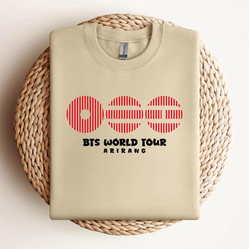 May include: A beige crewneck sweatshirt featuring a graphic of three red circular shapes with vertical stripes. Below the circles, the text reads BTS WORLD TOUR in a bold black font, followed by the word ARIRANG in a smaller black font.