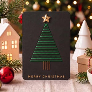 May include: A rectangular black greeting card featuring a stylised Christmas tree created with horizontal green thread stitching. A gold star is attached at the top, and four vertical gold lines form the trunk. The text MERRY CHRISTMAS is printed in gold.