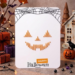 Può includere: Un biglietto d'auguri rettangolare bianco con un'illustrazione di ragnatela nera in alto e una faccia di zucca creata con punti di filo arancione verticali al centro. Il testo "HAPPY Halloween" è stampato nella parte inferiore.
