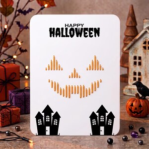 May include: A white rectangular card featuring the black text HAPPY HALLOWEEN at the top. Below the text is a jack-o'-lantern face created with vertical orange thread stitching. At the bottom are two black silhouettes of houses with windows.