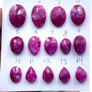 May include: A collection of fourteen faceted, oval and teardrop-shaped magenta gemstones arranged in three rows on a white card. Each stone is individually numbered from one to fourteen in handwritten ink above its respective position.