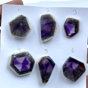 May include: Six faceted amethyst stones arranged on a white card, each numbered 1 through 6. The stones feature deep purple centers surrounded by a white border with black speckles. Each piece has a unique geometric shape and polished surface.