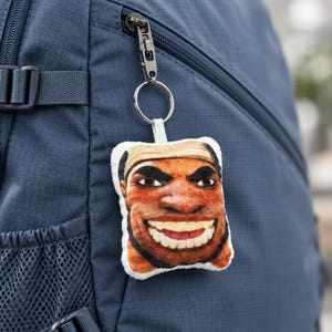 May include: A square-shaped plush keyring featuring a printed graphic of a person's face with a wide, toothy grin and a tan headband. The plush is attached to a silver-toned metal split ring and a black swivel clasp, hanging from a blue backpack.