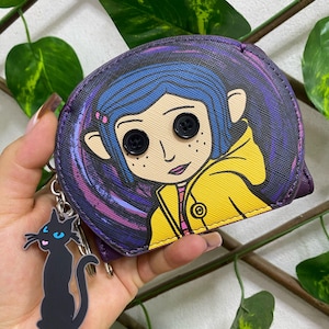 May include: A purple coin purse featuring a graphic of a character with blue hair, a yellow raincoat, and black button eyes. The purse includes a dangling black cat charm with blue eyes and a pink tongue attached to a silver metal keyring.