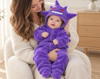 Puede incluir: Un mono de peluche morado para bebé con capucha que presenta detalles del personaje Gengar, incluyendo orejas puntiagudas, ojos rojos y una amplia sonrisa blanca con dientes. Prenda de manga larga con cremallera frontal.