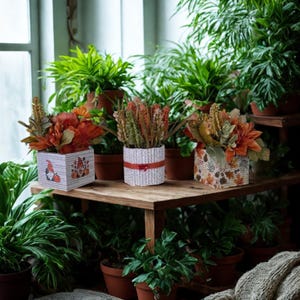 May include: Three square and cylindrical containers holding autumn-themed floral arrangements. The left container features gnome and pumpkin illustrations, the centre has a white textured wrap with a red ribbon, and the right displays a leaf pattern.