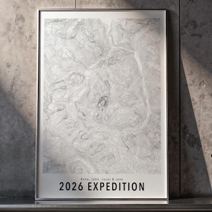 Könnte beinhalten: Ein gerahmtes vertikales Poster mit einer weißen topografischen Karte und grauen Höhenlinien. Unten steht der Text Anna, John, Lucas & Jane, gefolgt von 2026 EXPEDITION in fetten schwarzen Großbuchstaben. Der Druck befindet sich in einem silbernen Rahmen.